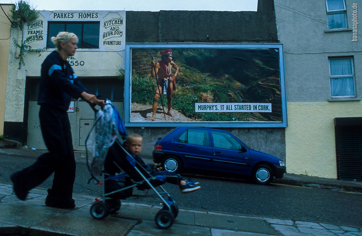 Frau mit Kinderwagen, im Hintergrund die Plakatwerbung It all startet in Cork